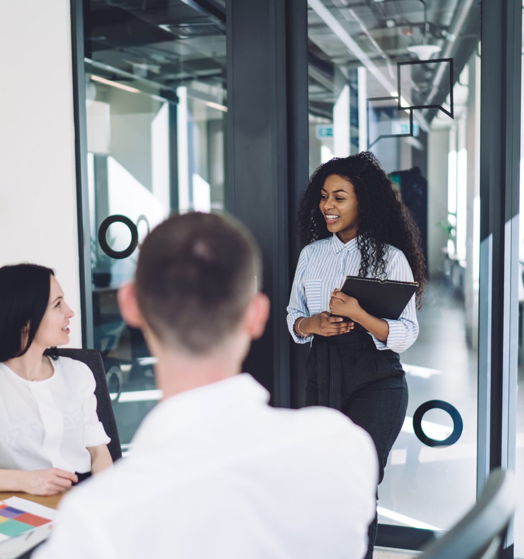 Black curly smiling lady with folder coming in startup company for meeting with hr service about job in modern conference room